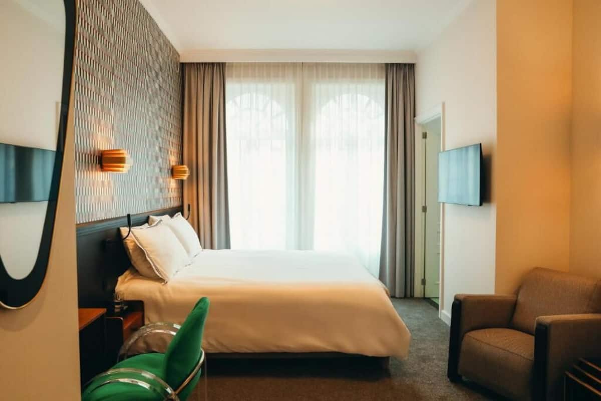 Ein Hotelzimmer mit einem Bett, einem grünen Sessel, einem braunen Ohrensessel, einem an der Wand montierten Fernseher und einem großen Fenster mit Vorhängen.