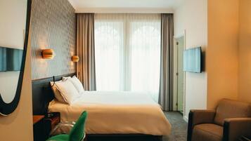 Habitación de hotel con una cama, una silla verde, un sillón marrón, un televisor montado en la pared y un ventanal con cortinas.