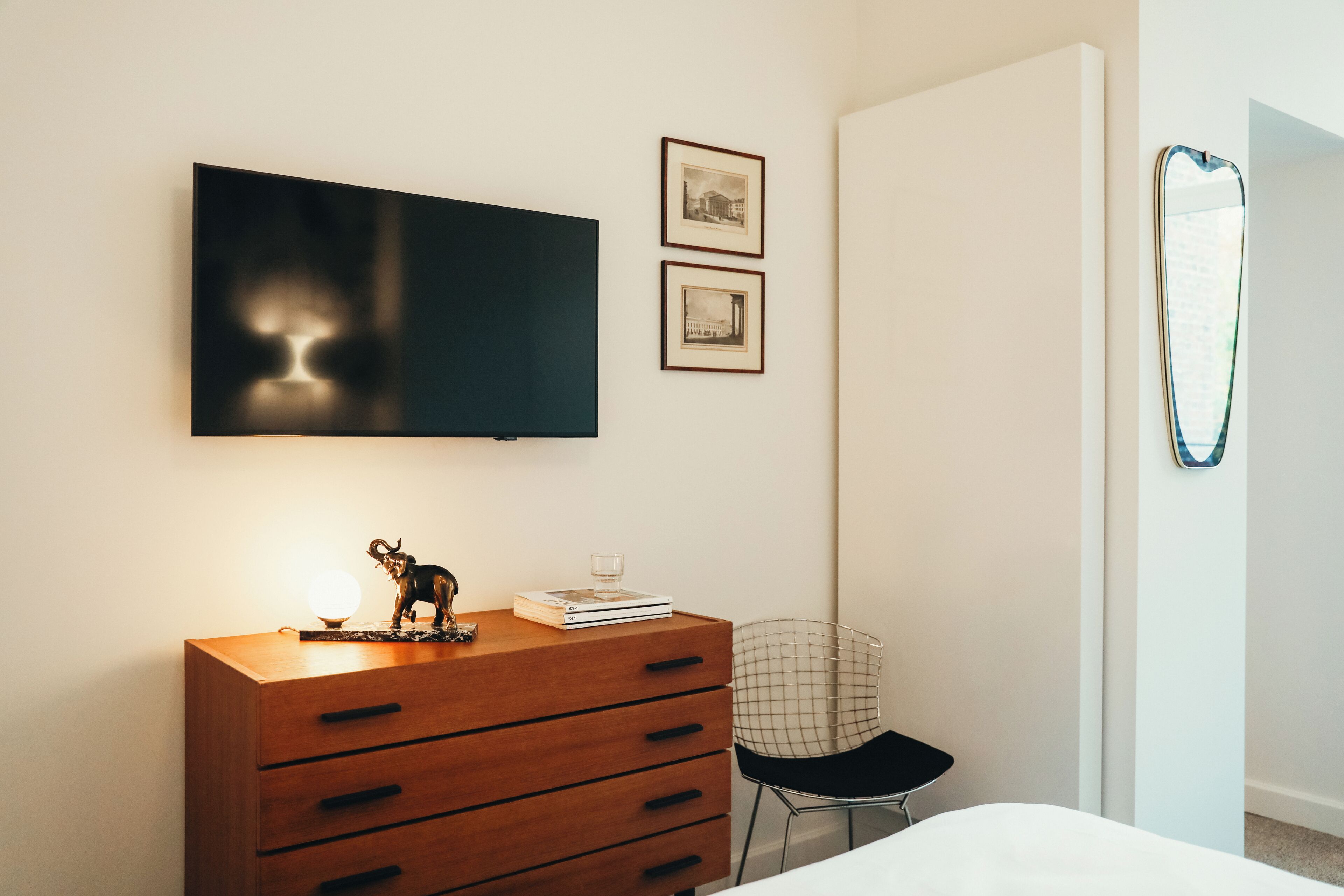 A bedroom with a wooden dresser, a bed, a chair, a TV mounted on the wall, and two framed pictures.