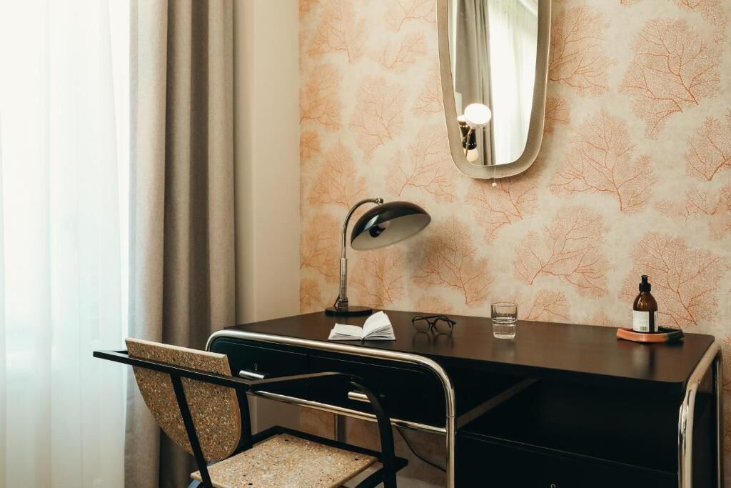 A hotel room with a desk, chair, mirror, lamp, book, glass of water, and a bottle on the desk.