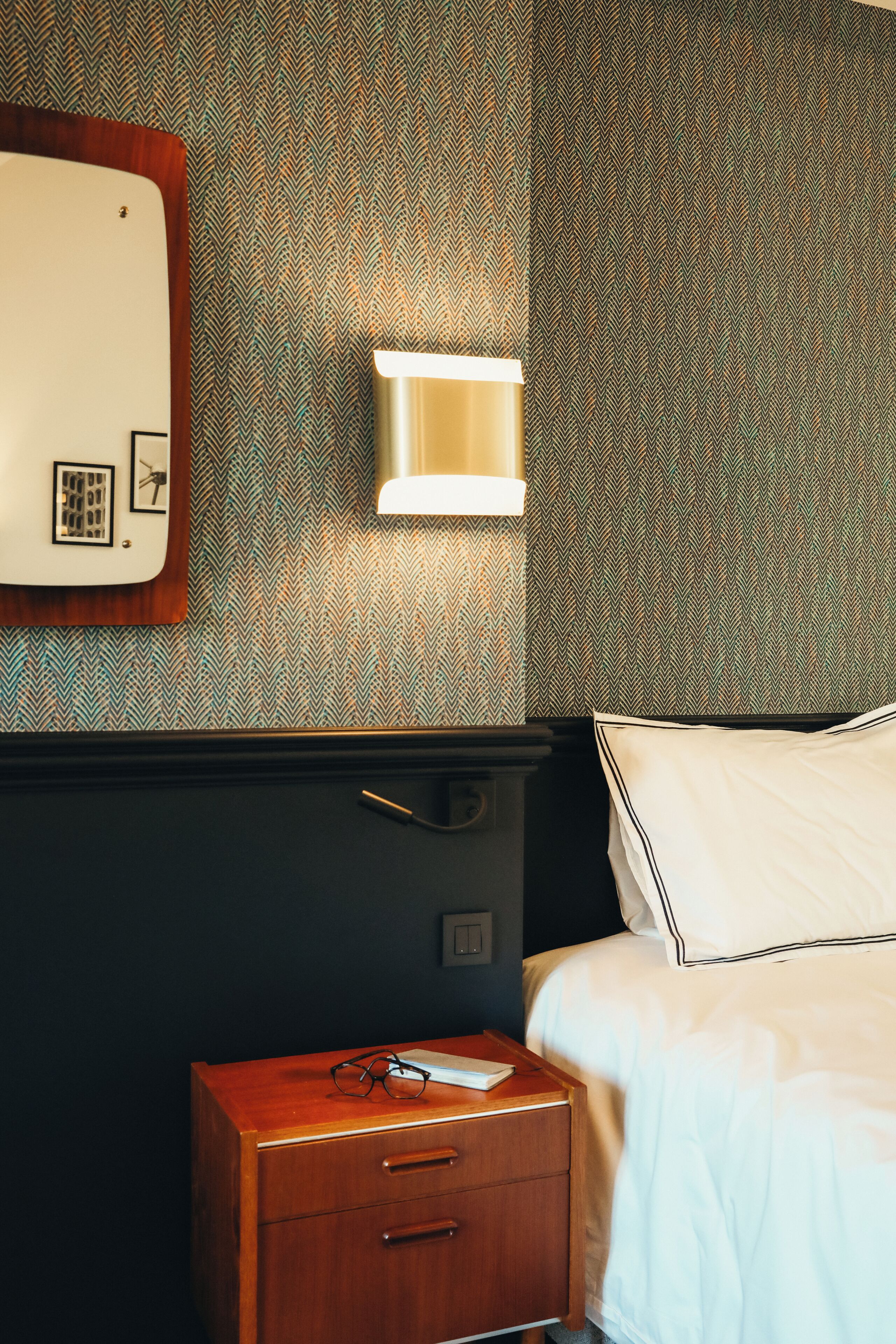 A hotel room with a wooden nightstand, a bedside table, a lamp, a bed with white bedding, and a patterned wallpaper.