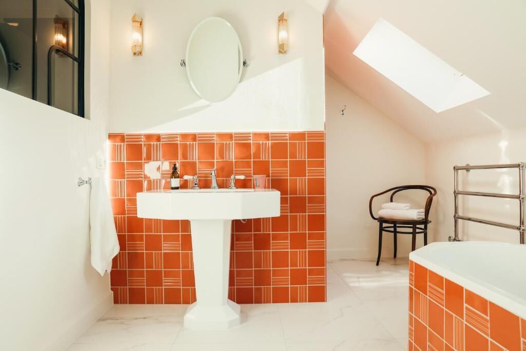 A bathroom with a white sink, orange tiled walls, a round mirror, and a skylight.