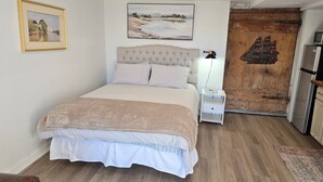 Un dormitorio con cama, mesita de noche, lámpara, un cuadro enmarco y puerta de madera con un mural de barco.