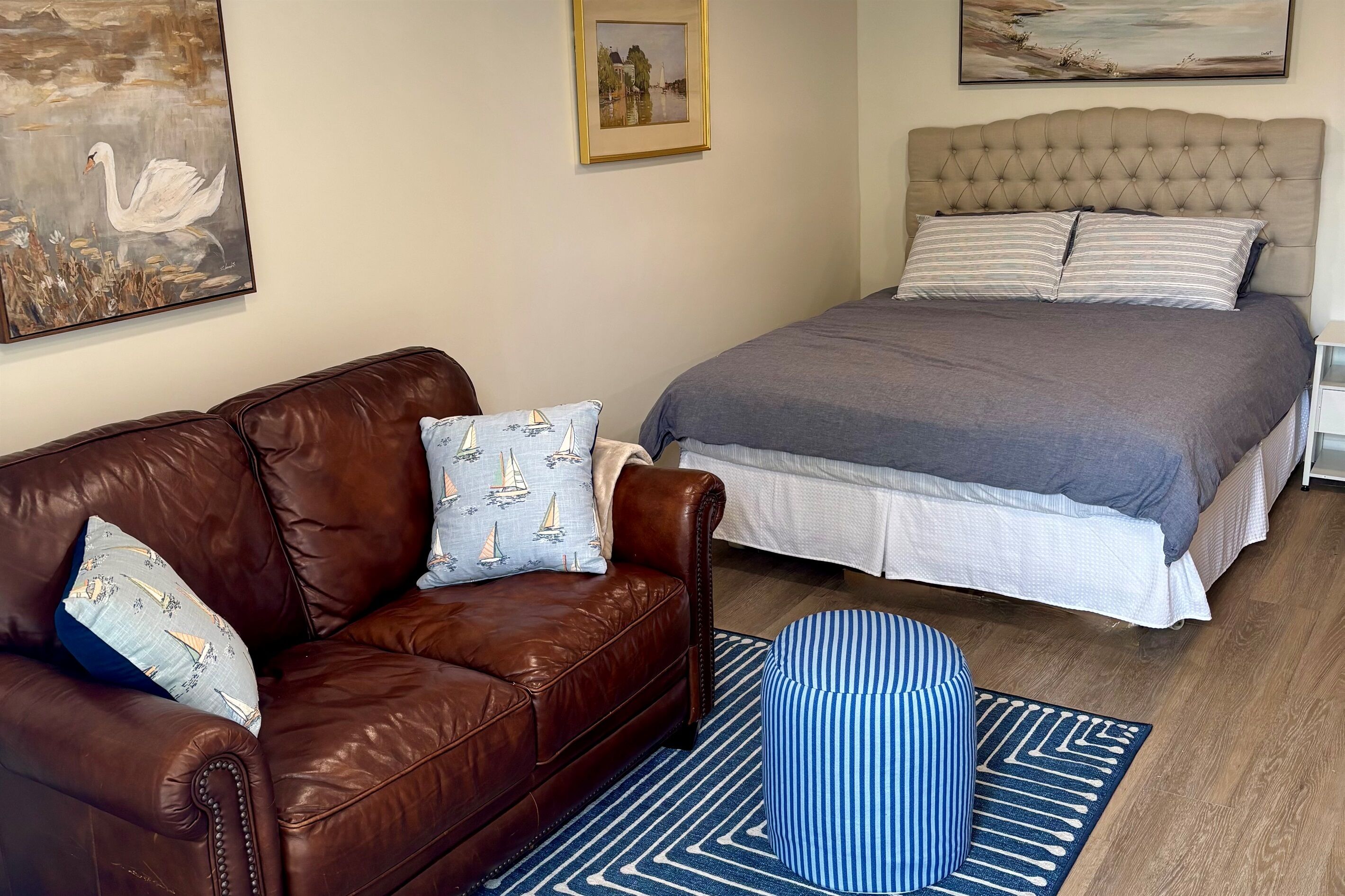 A bedroom with a brown leather sofa, a striped ottoman, a bed with a grey blanket, and two pillows.