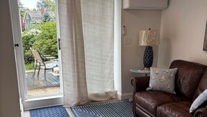 Una habitación con un sofá de cuero marrón, una mesita auxiliar con una lámpara, y vistas a un espacio exterior con una silla y vegetación.