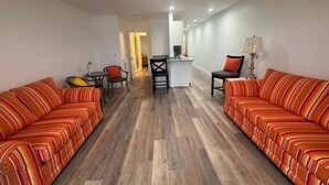 A living room with two striped sofas, a dining table with chairs, a kitchen area, and a hallway leading to another room. - #216 Polynesian - WALK 2 PIER PARK you and beach (Upper Grand Lagoon)