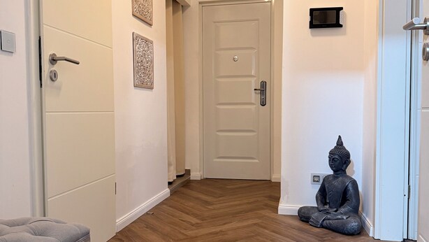 Un corridoio con pavimento in legno, una panca grigia, una statua di Buddha e una porta con pannello di sicurezza.