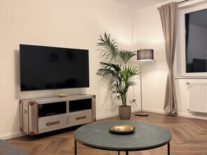 Ein Wohnzimmer mit einem Flachbildfernseher an einer Wandhalterung, einer Topfpflanze, einer Stehlampe, einem runden Tisch mit einer Schale und einem Fenster mit Vorhängen.