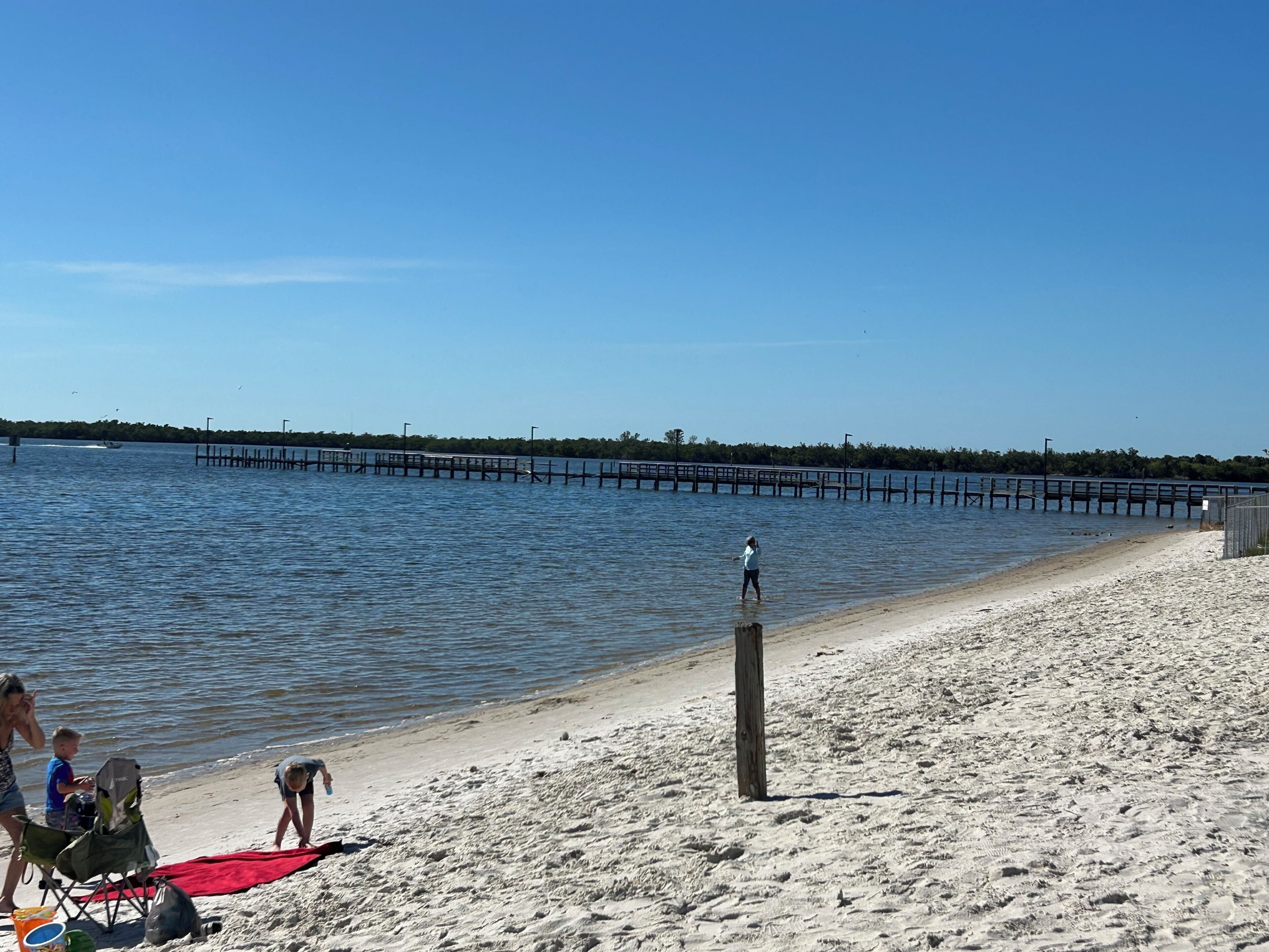 Une plage avec une jetée en bois s’avançant dans des eaux calmes, un rivage sablonneux, et quelques personnes et un chien à proximité.