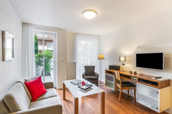 A hotel room with a sofa, a small table, a desk with a computer, a chair, a TV, and a window with curtains.