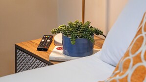 Una cama bien hecha con un almohadón blanco y uno estampado, un control remoto, una planta en maceta y una lámpara con pantalla de vidrio transparente sobre una mesita de noche de madera.