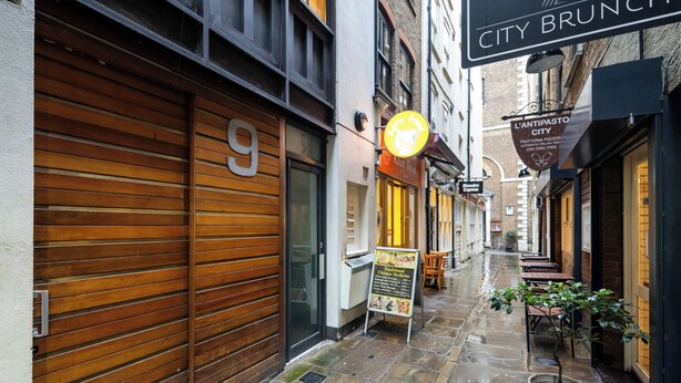 Una calle estrecha con contraventanas de madera y pavimento de piedra, un cartel que anuncia 'City Brunch' y una pizzería llamada 'L'ANTIPASTO CITY'.