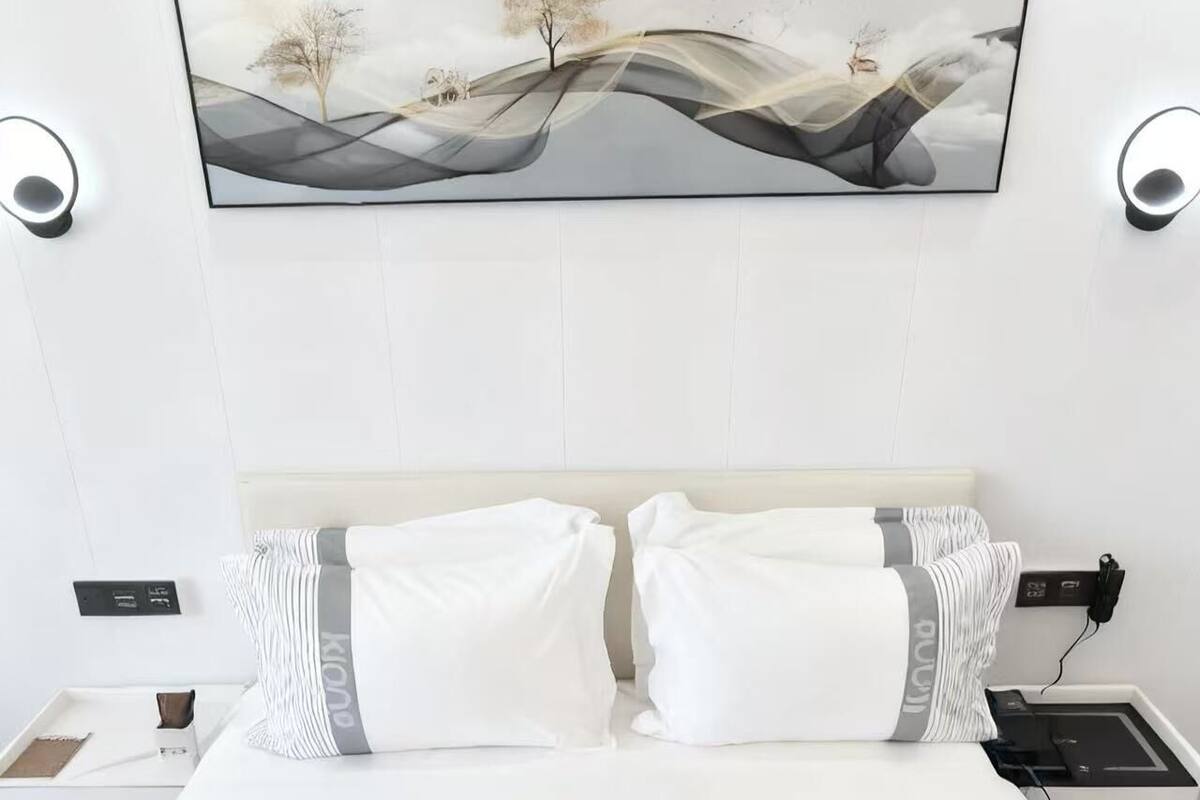 A modern hotel room with a large bed, white bedding, and a framed landscape painting above the bed. There are two bedside tables with lamps, a nightstand, and a power outlet on the wall.