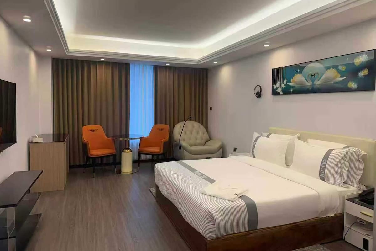 A hotel room with a bed, two chairs, a desk, a television, and a painting on the wall.