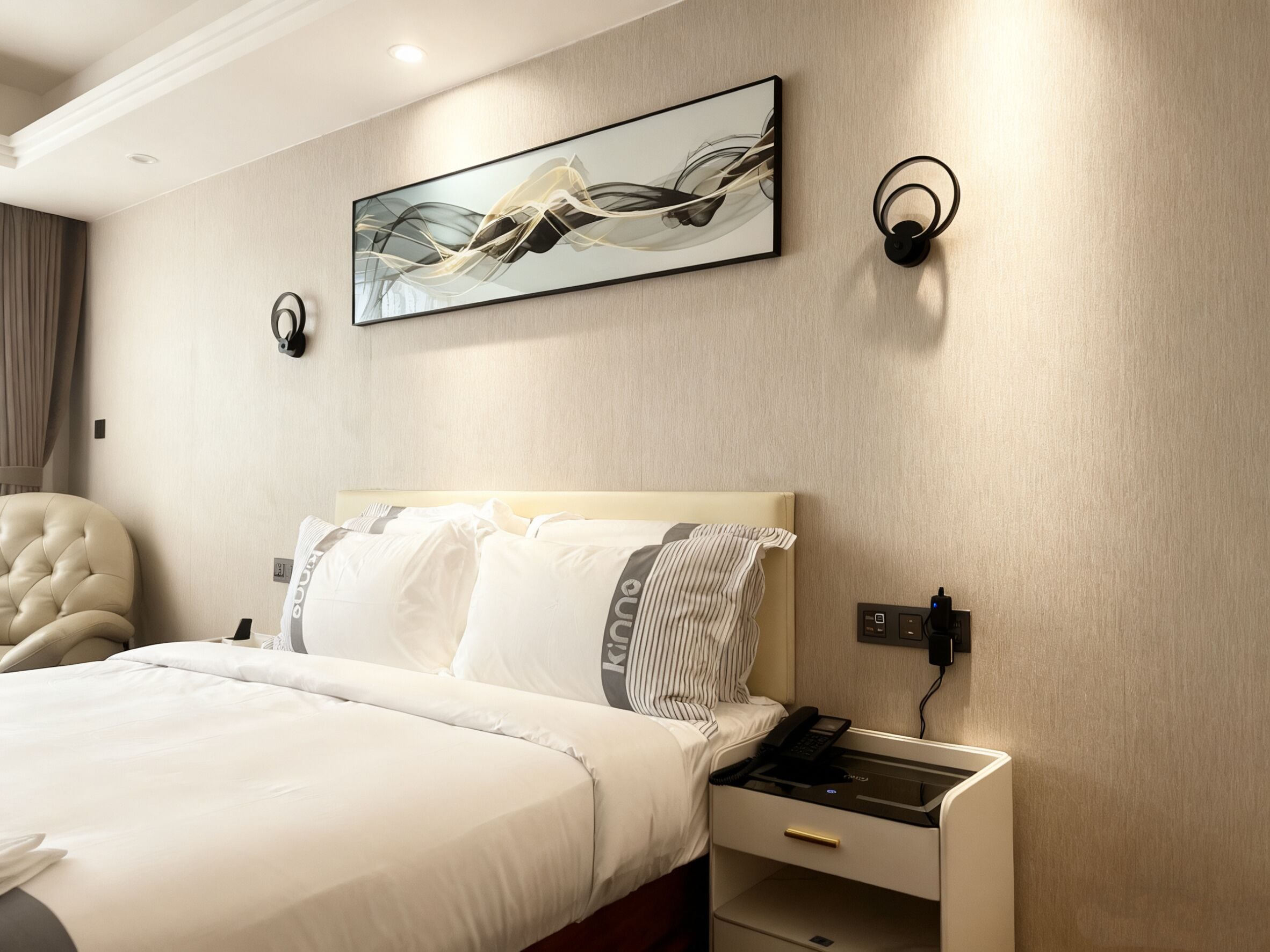 A hotel room with a large bed, a bedside table with a phone, a wall-mounted light, and a painting above the bed.