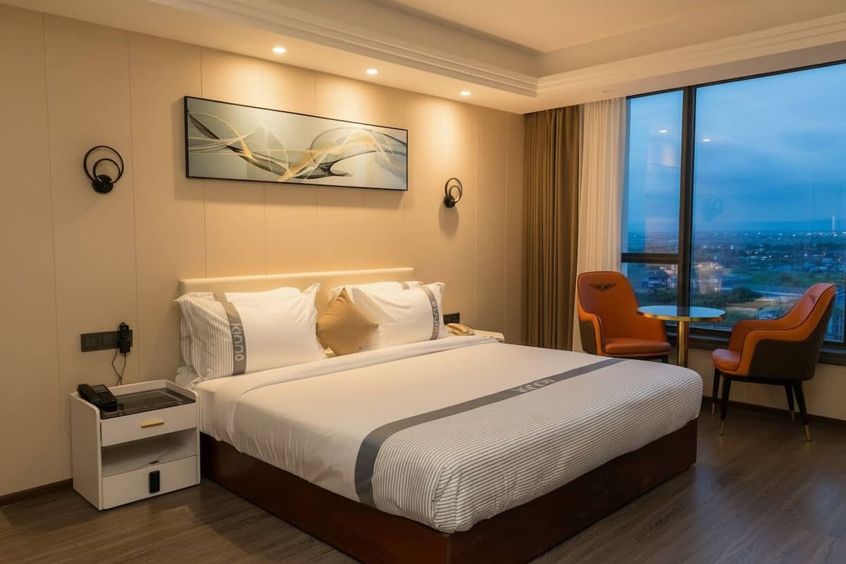 A modern hotel room with a large bed, a desk with a telephone, and a view of the city through large windows.