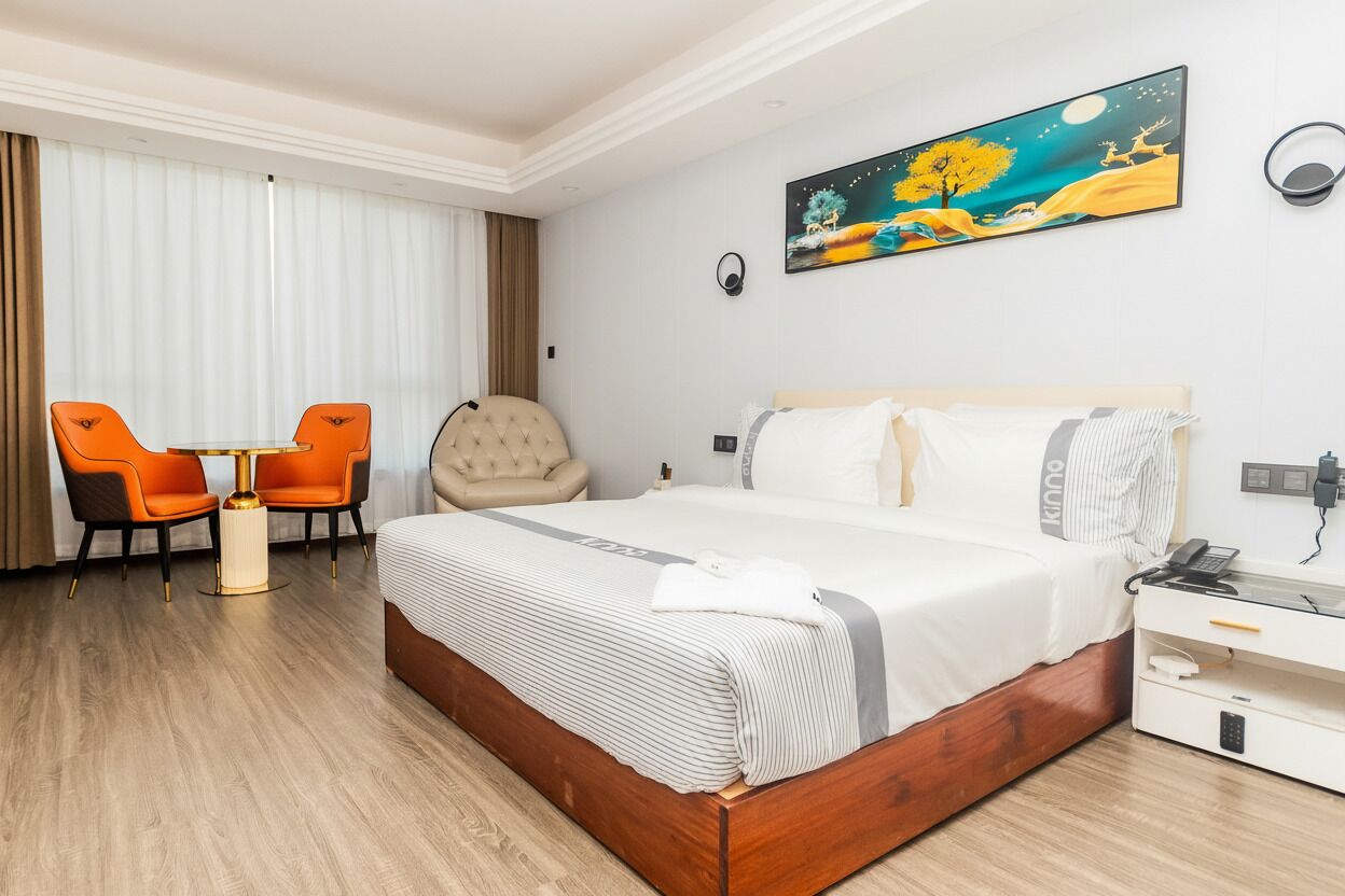 A hotel room with a large bed, a bedside table with a phone, a chair, a small table, and a painting on the wall.