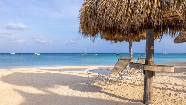 Une plage avec un parasol en chaume, deux chaises longues et un poteau en bois portant une pancarte.