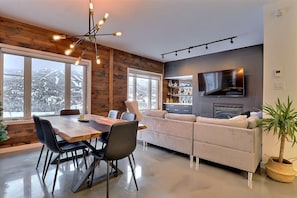 A modern living room with a large window, a dining area with a wooden table, and a grey sofa. - Condos Monts-Valin -153, rue du Massif (Valinouët) (Le Fjord-du-Saguenay Regional County Municipality)