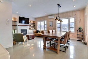 A spacious living area with a wooden dining table, comfortable seating, a fireplace, and large windows offering a view of the outdoors. - Condos Monts-Valin - 47, rue du Sommet (Valinouët) (Saint-David-de-Falardeau)