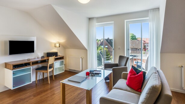 Una sala de estar moderna con una televisión de pantalla plana, un escritorio con una silla, un sofá con cojines rojos y un ventanal con vistas al exterior.