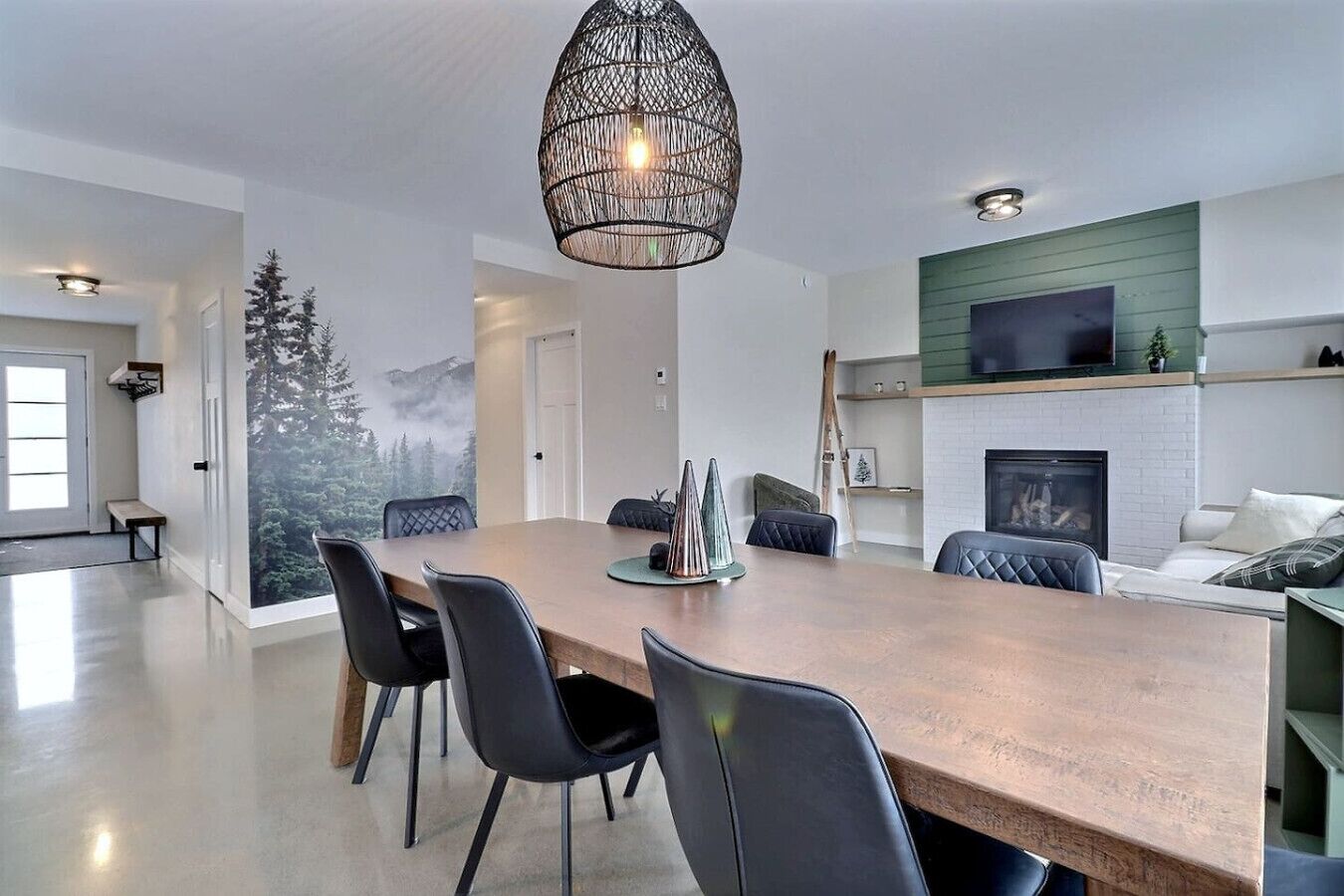 A modern living room with a large wooden dining table, black chairs, a fireplace, and a green accent wall.