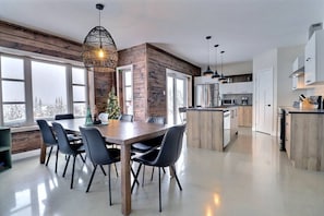 A modern kitchen with a central island, wooden accents, and a large dining area with a chandelier. - Condos Monts-Valin - 27, rue du Sommet (Valinouët) (Saint-David-de-Falardeau)