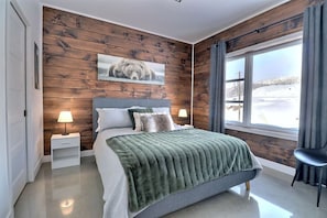 A modern bedroom with a wooden wall panel, a large bed with a green blanket, two bedside lamps, and a window with a view of a snowy landscape. - Condos Monts-Valin - 49, rue du Sommet (Valinouët) (Saint-David-de-Falardeau)