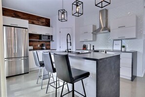 A modern kitchen with stainless steel appliances, a central island with bar stools, and pendant lighting. - Condos Monts-Valin - 49, rue du Sommet (Valinouët) (Saint-David-de-Falardeau)