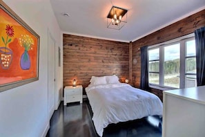 A bedroom with a wooden wall, a bed, a nightstand, a painting, and a window with a view. - Condos Monts-Valin- 213, rue du Massif (Valinouët) (Saint-David-de-Falardeau)