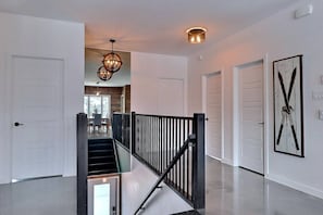 A spacious hallway with a black railing, white doors, and a chandelier. - Condos Monts-Valin- 165, rue du Massif (Valinouët) (Le Fjord-du-Saguenay Regional County Municipality)