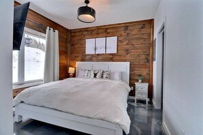 A bedroom with a white bed, wooden walls, a window with curtains, and a bedside table with a lamp. - Condos Monts-Valin- 165, rue du Massif (Valinouët) (Le Fjord-du-Saguenay Regional County Municipality)