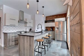 A modern kitchen with a central island, bar stools, stainless steel appliances, and pendant lighting. - Condos Monts-Valin- 165, rue du Massif (Valinouët) (Le Fjord-du-Saguenay Regional County Municipality)