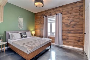 A bedroom with a wooden wall, a bed with a grey headboard, a macrame wall hanging, and a window with curtains. - Condos Monts-Valin- 165, rue du Massif (Valinouët) (Le Fjord-du-Saguenay Regional County Municipality)