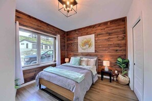 A bedroom with a wooden wall, a bed, a window, and a nightstand with a lamp. - Condos Monts-Valin -151, rue du Massif (Valinouët) (Saint-David-de-Falardeau)