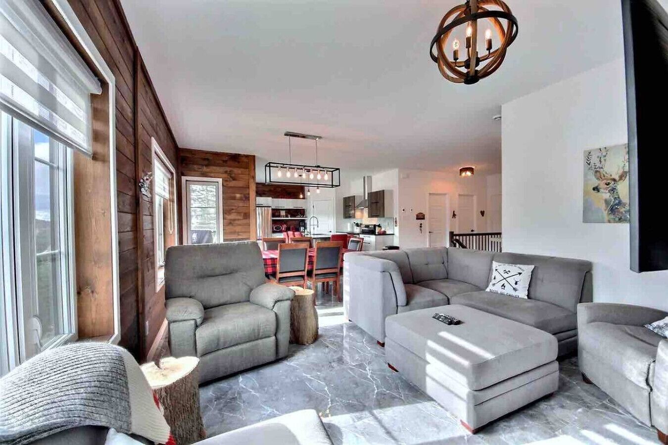 A modern living room with a grey sectional sofa, a grey armchair, and a coffee table. There is a wooden accent wall and a chandelier.