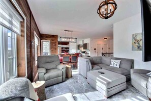 A modern living room with a grey sectional sofa, a grey armchair, and a coffee table. There is a wooden accent wall and a chandelier. - Condos Monts-Valin -149, rue du Massif (Valinouët) (Saint-David-de-Falardeau)