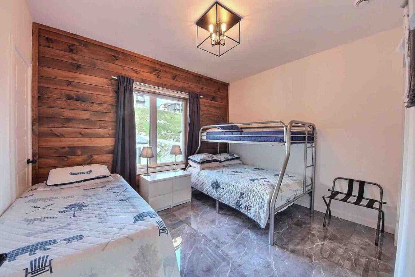 Une pièce aux murs lambrés, avec un lit superposé, un lit simple, une fenêtre avec des rideaux, une table de chevet et une chaise.