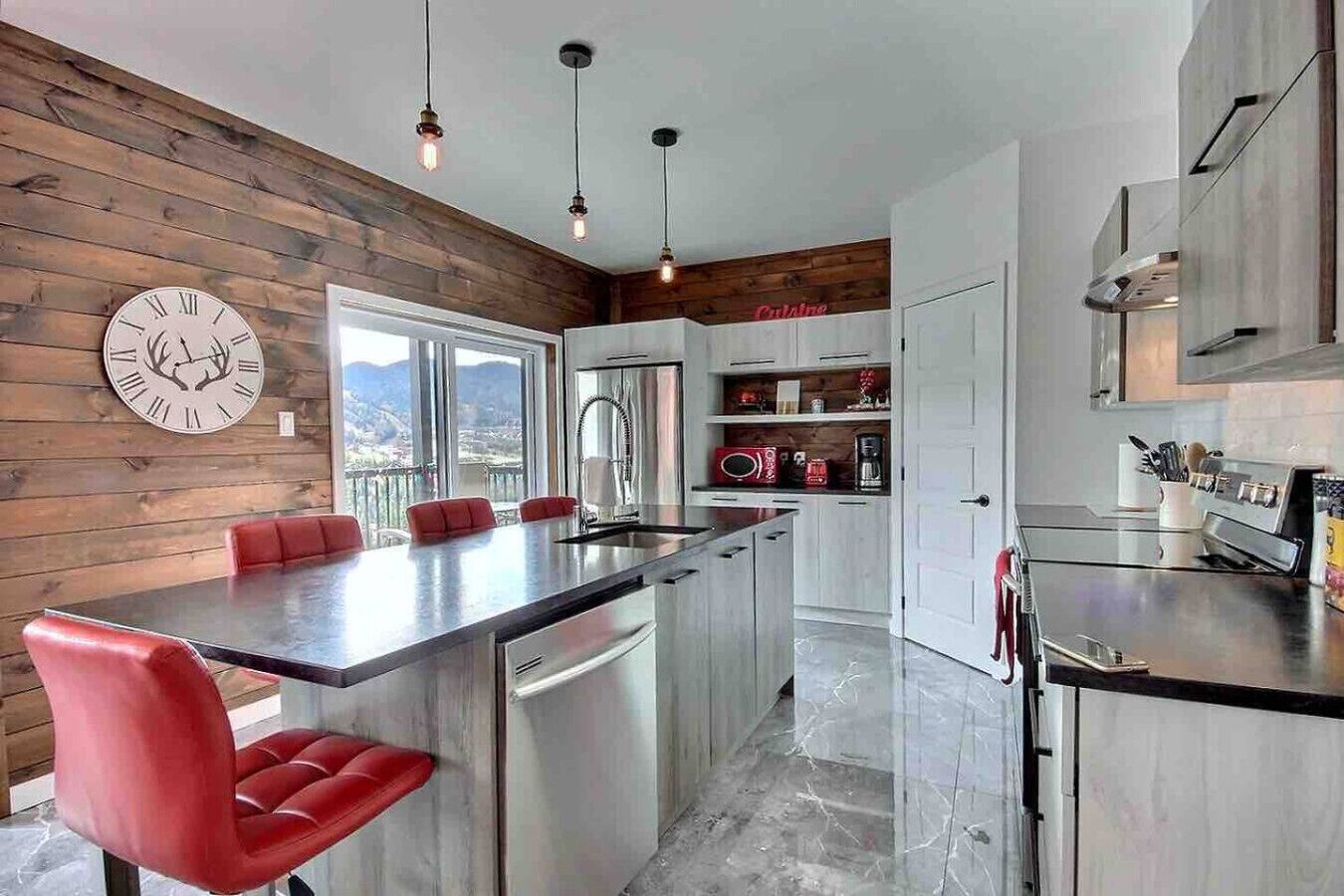 A modern kitchen with a central island, red chairs, and a wooden wall with a clock.