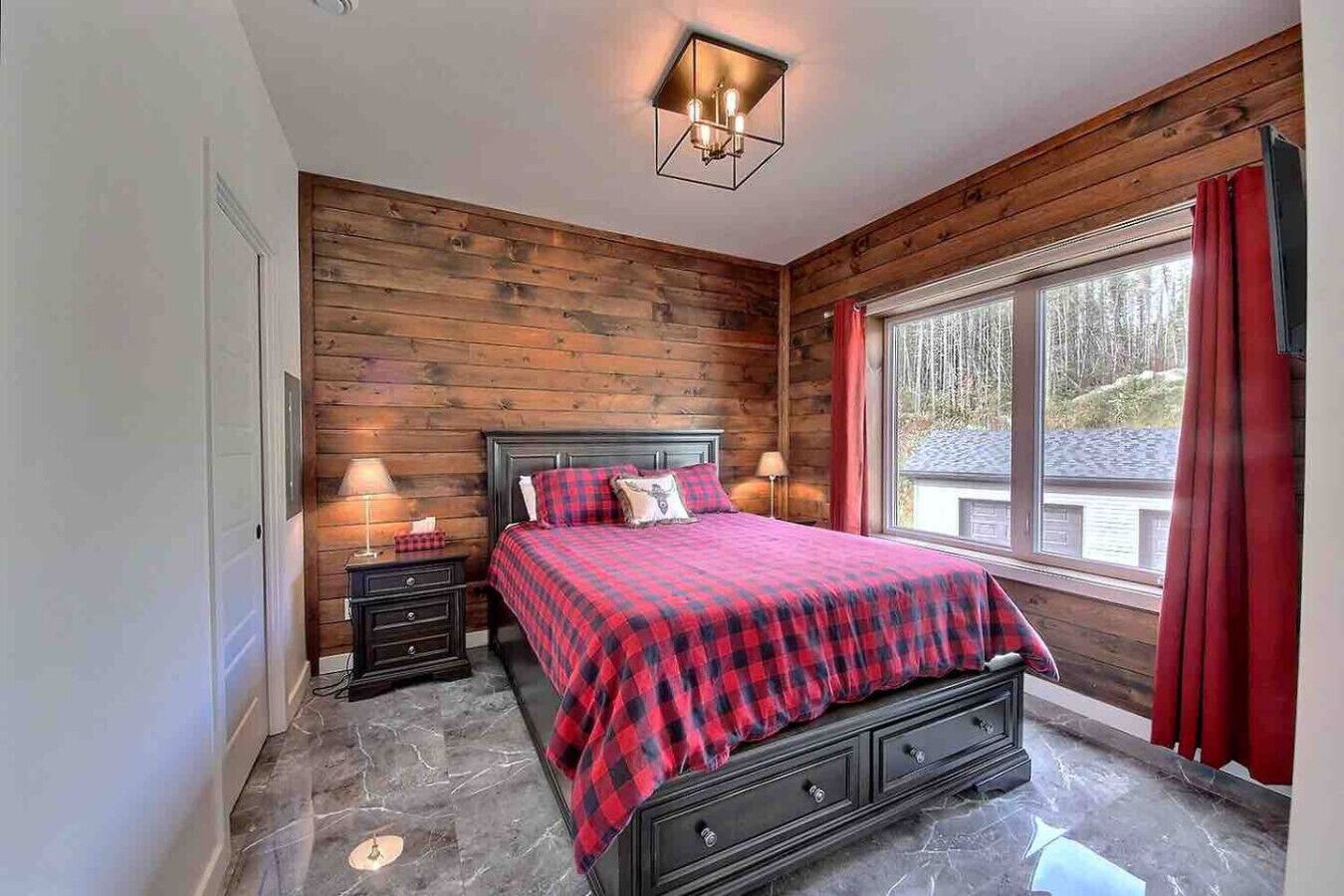 Une chambre avec un mur en bois, un lit recouvert d’une couette à carreaux rouges et noirs, deux tables de chevet avec des lampes, et une fenêtre avec des rideaux rouges.