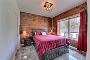 A bedroom with a wooden wall, a bed with a red and black checkered bedspread, two bedside tables with lamps, and a window with red curtains. - Condos Monts-Valin -149, rue du Massif (Valinouët) (Saint-David-de-Falardeau)