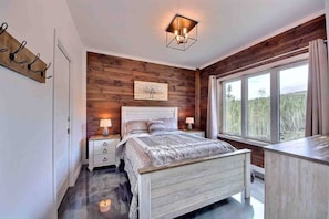 A bedroom with a wooden wall, a bed with a white headboard, two bedside tables with lamps, and a window with a view of trees. - Condos Monts-Valin- 203, rue du Massif (Valinouët) (Saint-David-de-Falardeau)