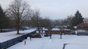Área externa coberta de neve, com cercado, árvores e um edifício com telhado vermelho.