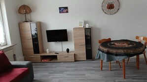 Uma sala de estar com televisão, mobiliário de madeira, uma mesa de jantar com toalha de mesa estampada e um sofá vermelho.