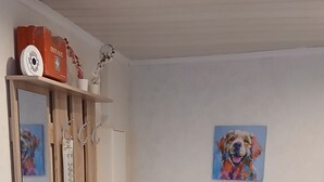 Um corredor com piso de madeira, uma chaleira vermelha sobre um armário de madeira, um armário branco, um termômetro fixado na parede e um quadro com a imagem de um cachorro.