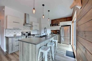 A modern kitchen with a central island, bar stools, stainless steel appliances, and pendant lighting. - Condos Monts-Valin -161, rue du Massif (Valinouët) (Saint-David-de-Falardeau)