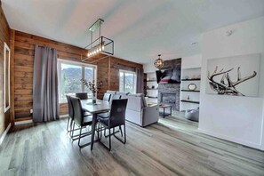 A modern living room with a fireplace, a dining area with a table and chairs, and a large window with curtains. - Condos Monts-Valin -161, rue du Massif (Valinouët) (Saint-David-de-Falardeau)