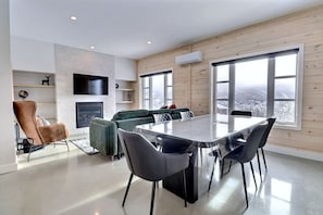 A modern living room with a fireplace, a dining table, and large windows offering a view of a snowy landscape. - Condos Monts-Valin - 29, rue du Sommet (Valinouët) (Saint-David-de-Falardeau)