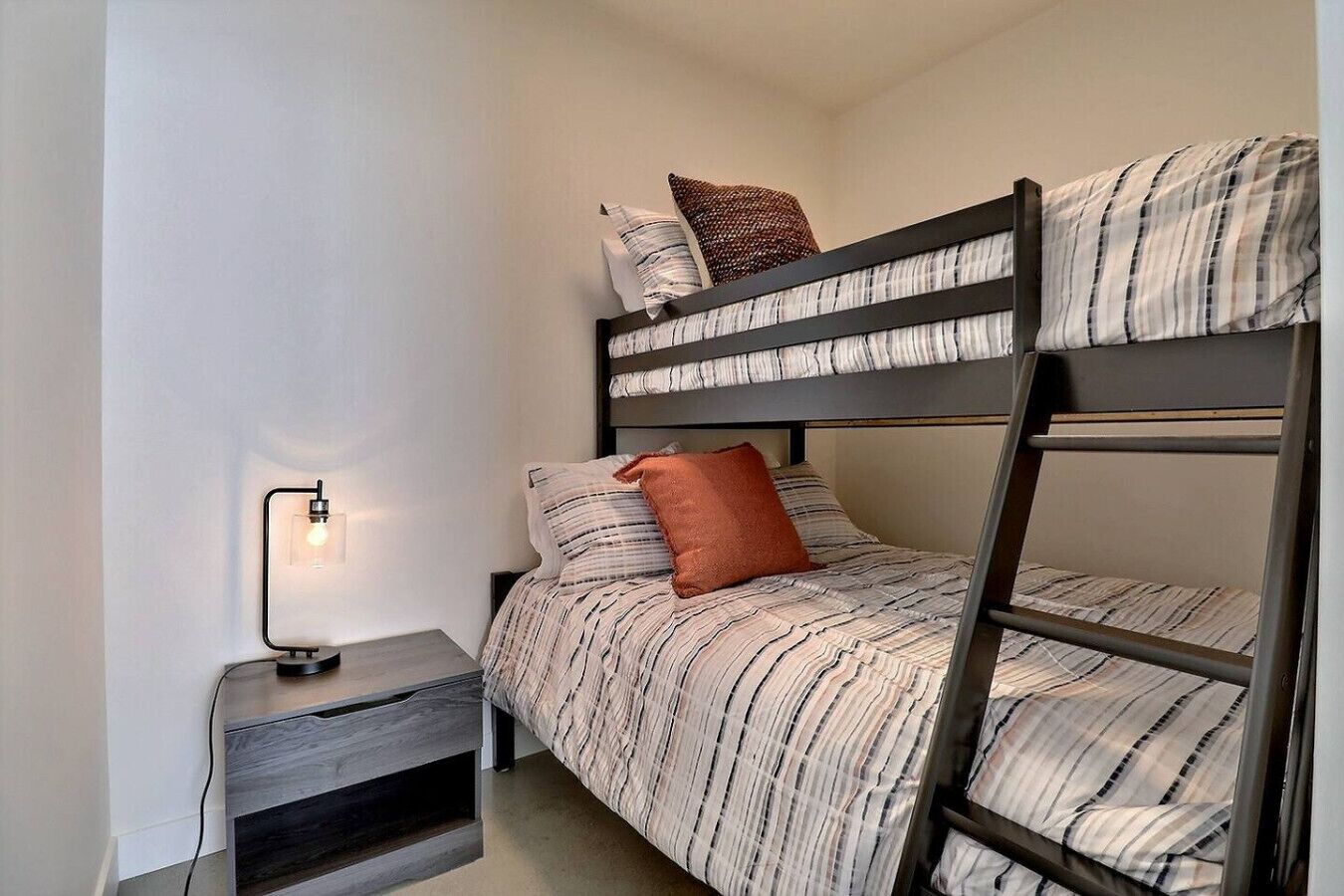 A bunk bed with striped bedding, a bedside table with a lamp, and a wall-mounted shelf.