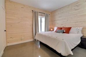 A bedroom with a wooden paneled wall, a bed with white bedding, a nightstand, and a window with curtains. - Condos Monts-Valin - 29, rue du Sommet (Valinouët) (Saint-David-de-Falardeau)
